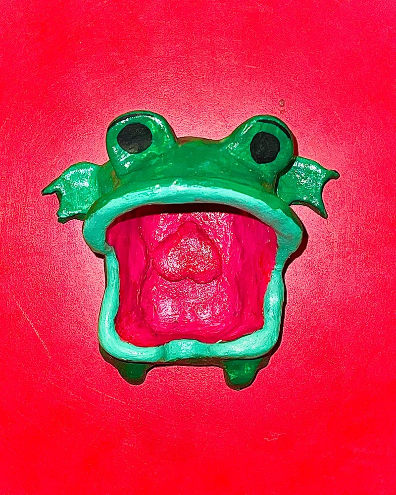 Handmade green frog pot with a wide-open pink mouth, on a red surface